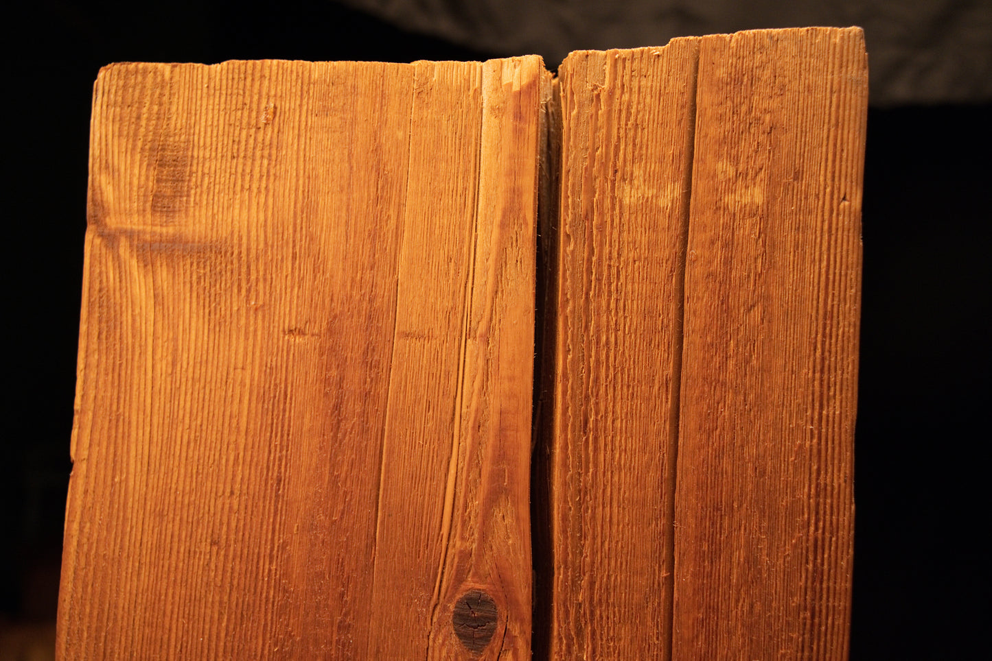 Macro detail of a narrow vertical surface split intersecting a dark organic knot near the upper face of a wooden beam. 