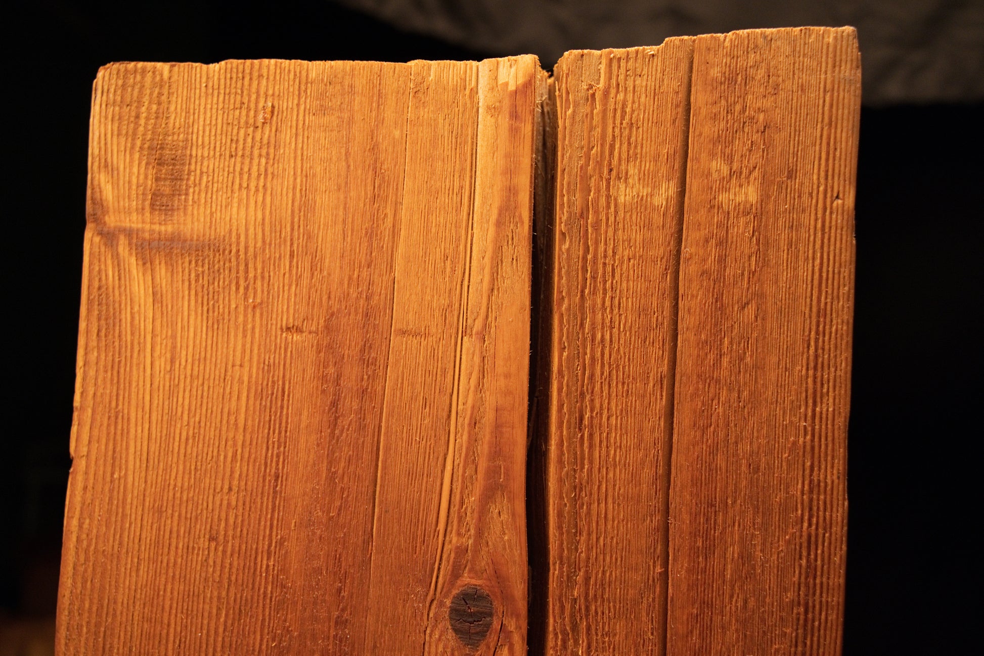 Macro detail of a narrow vertical surface split intersecting a dark organic knot near the upper face of a wooden beam. 