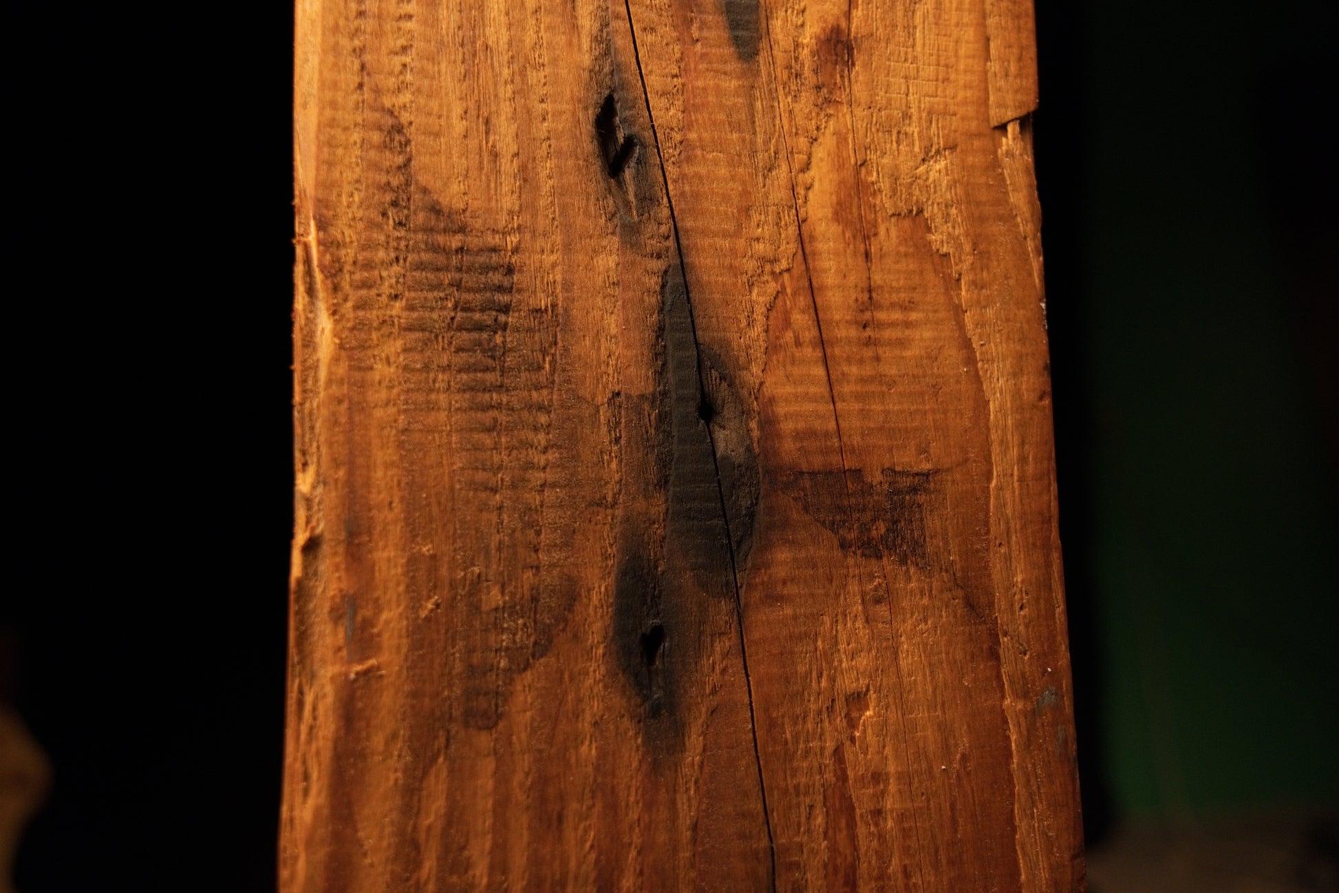 Close detail of a diagonal surface split intersecting dark knot cavities on the textured vertical face of a wooden monolith. 