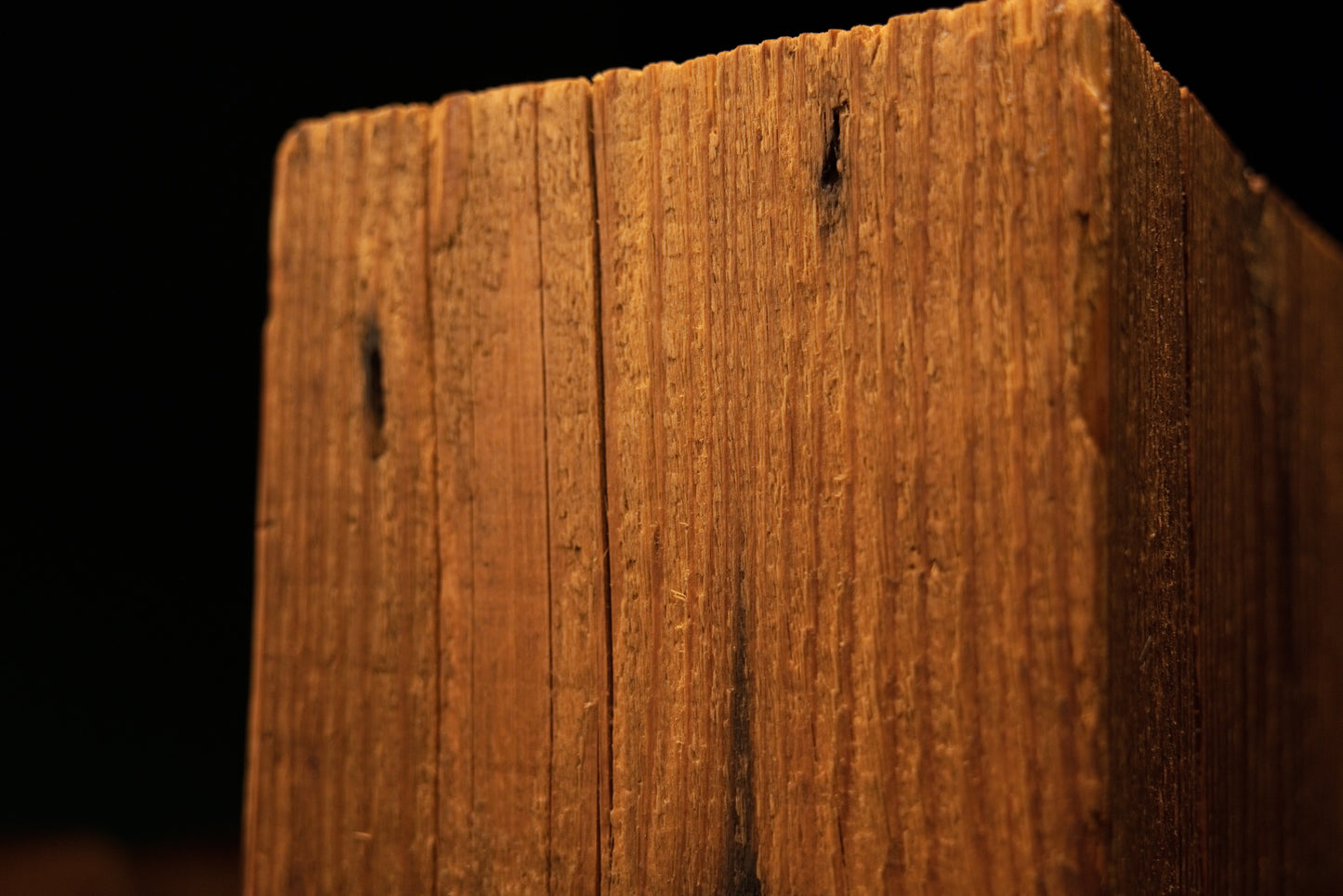 Close-up of the upper corner section on a timber post displaying linear grain and small dark pinholes near the edge. 
