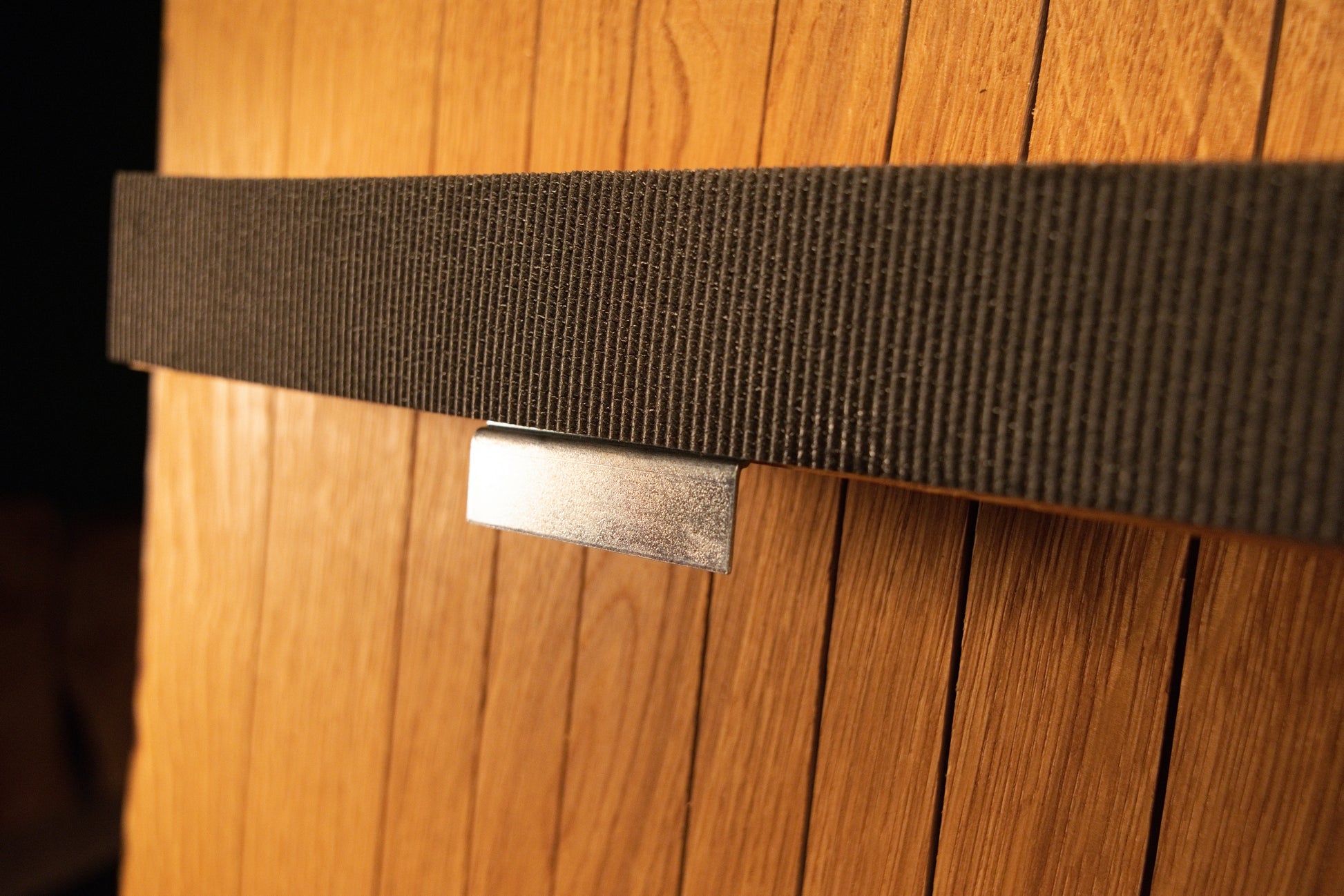 Detailed close-up view of a polished metal mounting clip securing the textured fabric strap against the vertical solid oak slats.