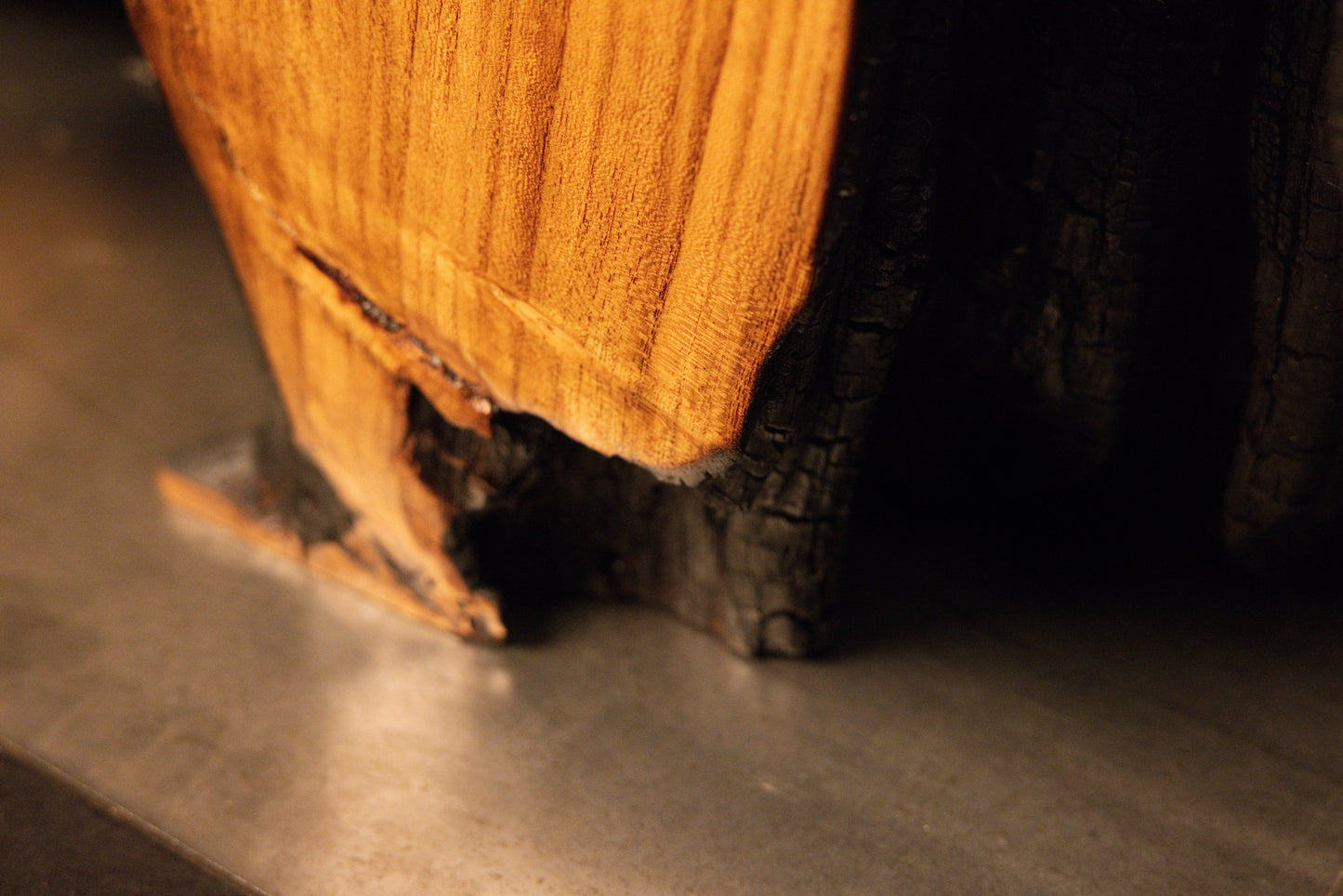 Foundation area detail where a warm golden wood face converges with the dark charred core and metallic floor base.  